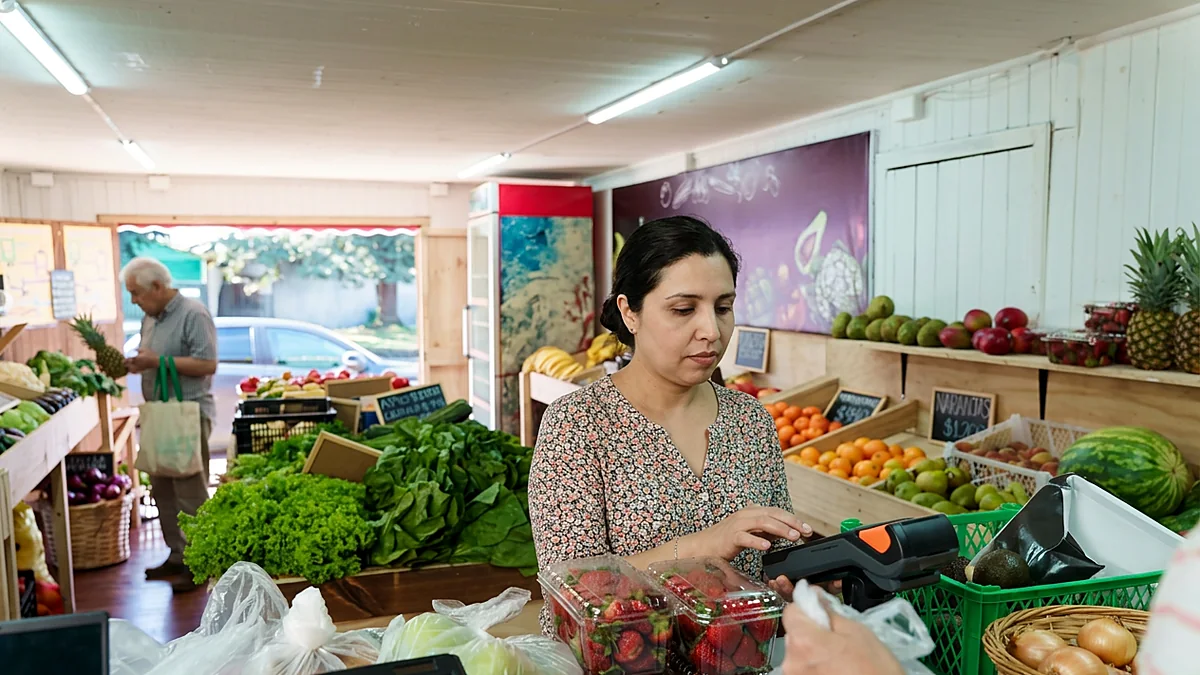 Cajera de una verdulería de barrio cobra con una máquina de tarjetas, en un almacén con frutas y verduras